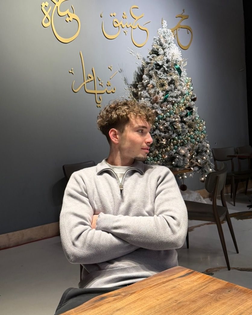 Yamen sitting at a wooden café table with his arms crossed, gold Arabic calligraphy on the dark wall behind him.